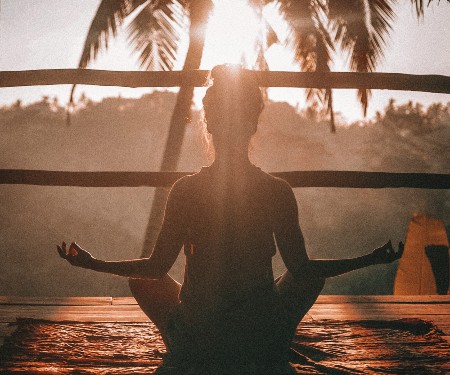 Woman sitting down in a yoga position, looking at palm trees as the sun sets